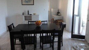 Un coin repas avec une table, des chaises et un saladier rempli de fruits. Au fond, on aperçoit une cuisine équipée d’un réfrigérateur et d’une machine à café.