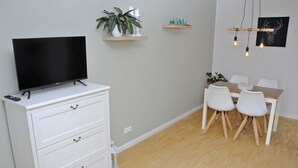 Ein modernes Wohnzimmer mit einem Flachbildfernseher auf einer weißen Kommode, einem Esstisch mit vier Stühlen und einer Wandleuchte.