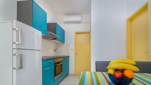 Cocina compacta con gabinetes color turquesa, un refrigerador blanco y una puerta amarilla. Se ve una mesa con rayas de colores y un frutero con plátanos, manzanas y naranjas.