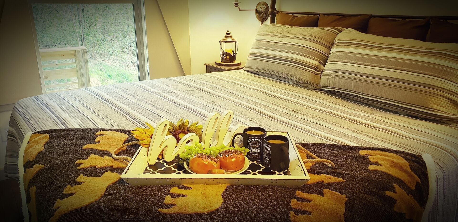 Una cama bien tendida con ropa de cama a rayas, una bandeja con alimentos para el desayuno y una mesita de noche con una lámpara.