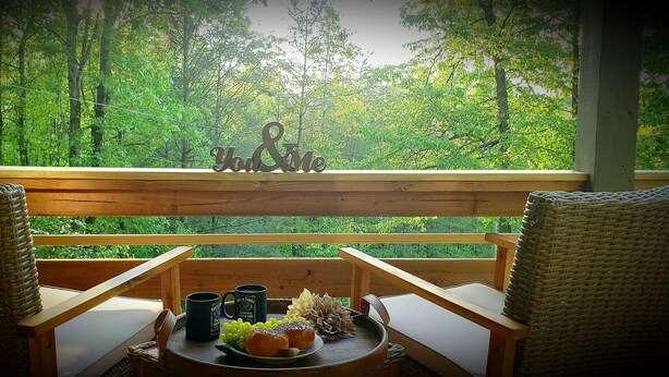A balcony with wicker chairs, a table with a plate of food, two mugs, and a decorative sign. - Happy Camper 1 Bedroom Cottage by RedAwning (Sevierville)