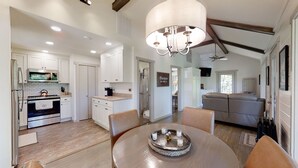 A spacious kitchen with white cabinets, a stainless steel refrigerator, and a kettle on the stove. There is a dining area with a round table and chairs, and a chandelier hanging above. - Happy Camper 1 Bedroom Cottage by RedAwning (Sevierville)