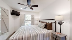 Un dormitorio con cama, ventilador de techo, lámpara de noche y televisor montado en la pared.