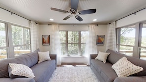Una habitación con dos sofás grises, un ventilador de techo y ventanas amplias con cortinas.