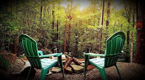 Two green Adirondack chairs facing a campfire in a forested area.