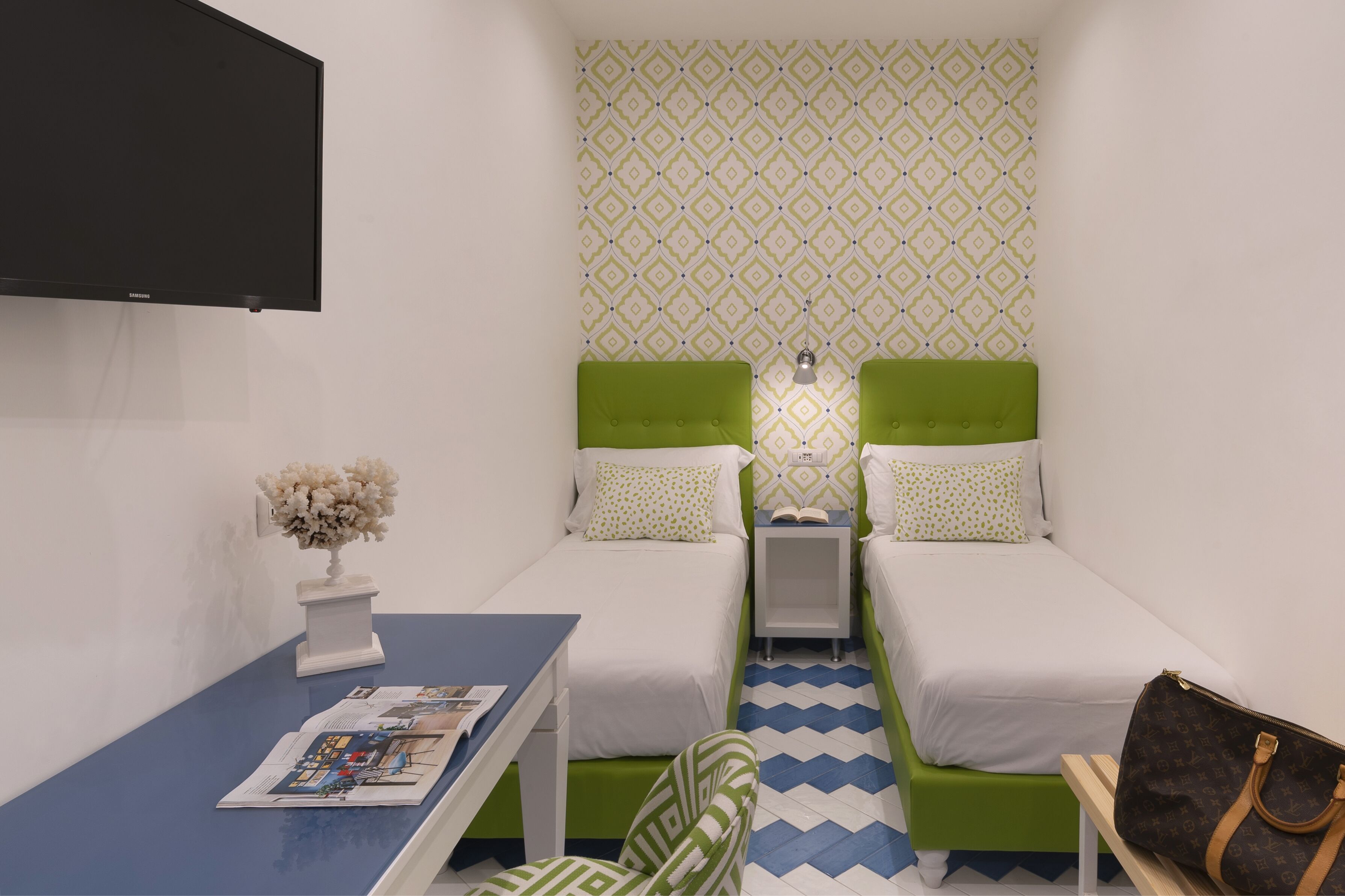 A small, well-lit room with two single beds, a desk with a chair, a flat-screen TV, and a patterned wall.