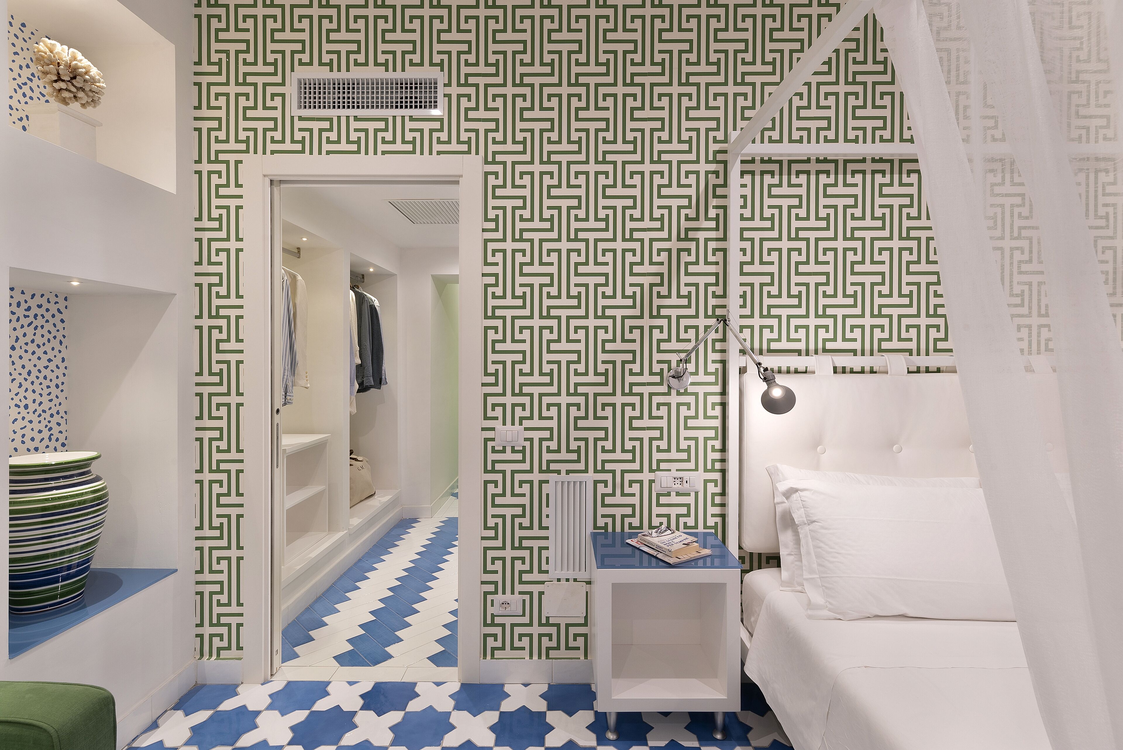 A hotel room with a geometric patterned wallpaper, a white bed, a bedside table with a lamp, and a blue and white tiled floor.
