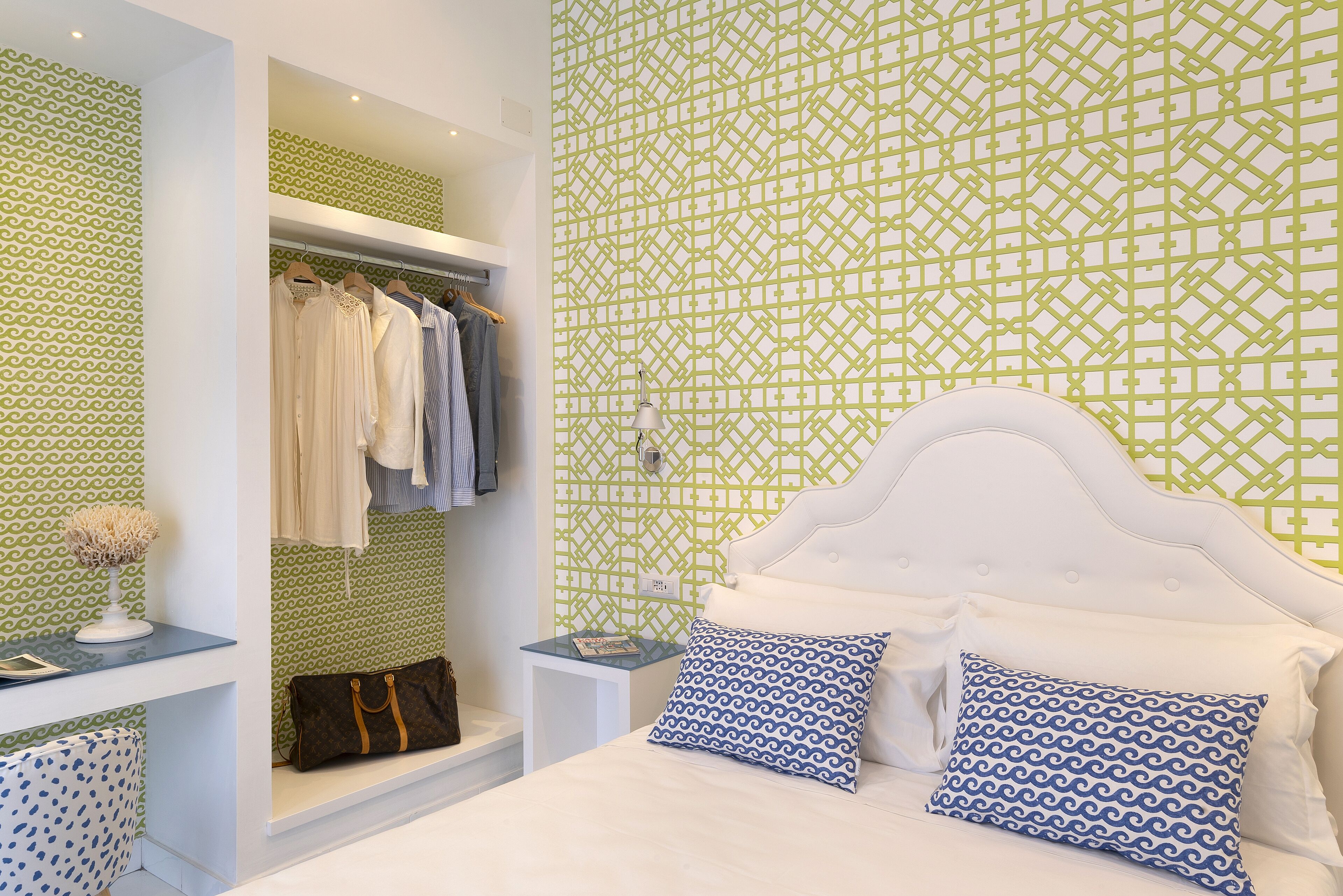 Un dormitorio con pared verde y blanca estampada, una cama blanca con dos almohadas, una mesita de noche con lámpara y un armario con ropa.