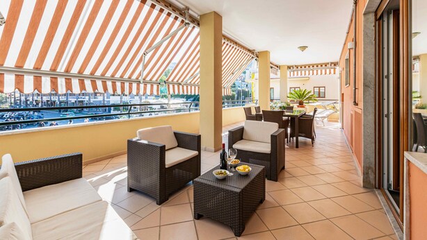 Un balcón con muebles de mimbre, una mesa con botella y fruta, y vista a un puerto deportivo.