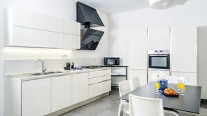 Una cocina moderna con armarios blancos, una estufa a gas, microondas y horno empotrado. Cuenta con una mesa de comedor con sillas y una mesa para desayunar con cruasanes y jugo de naranja.