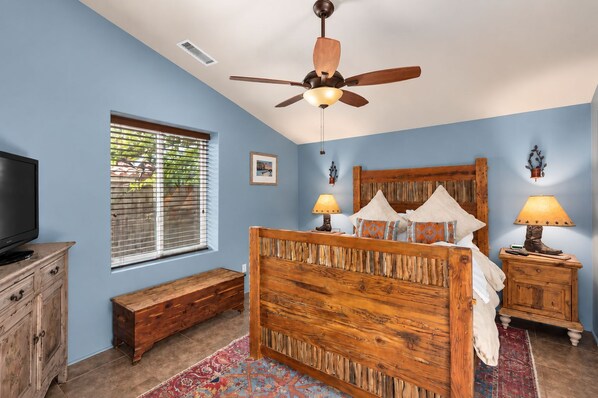 A bedroom with a wooden bed, a ceiling fan, a television, a window with blinds, and a nightstand with a lamp.