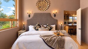 A bedroom with a large bed, a headboard, bedside lamps, a wall-mounted light fixture, and a view of the kitchen. - Meadow Lark Trail 3BD 2BA W/ Jacuzzi in THE best location (Sedona)