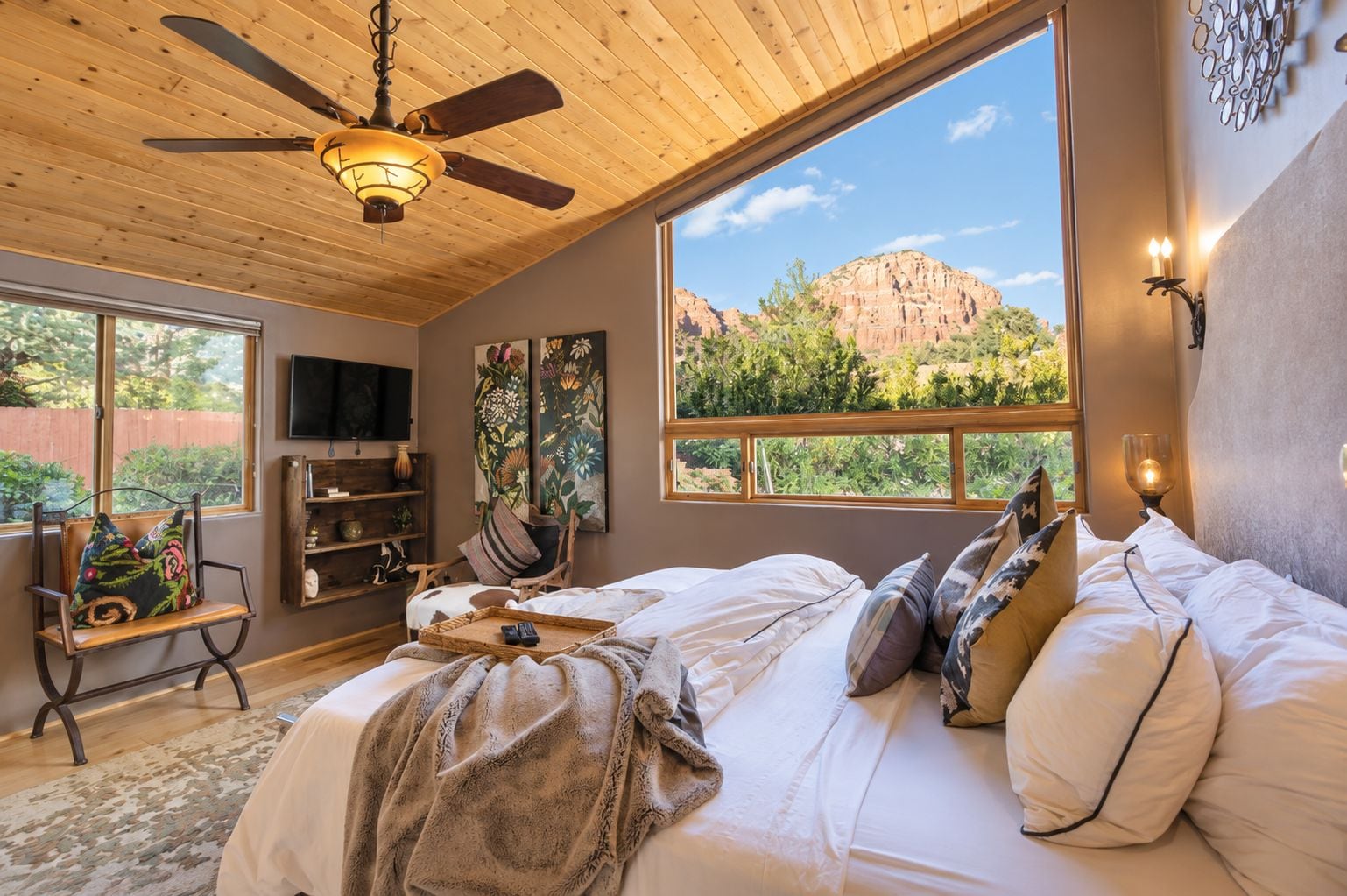 A bedroom with a large bed, a ceiling fan, a TV, and a view of red rock formations.