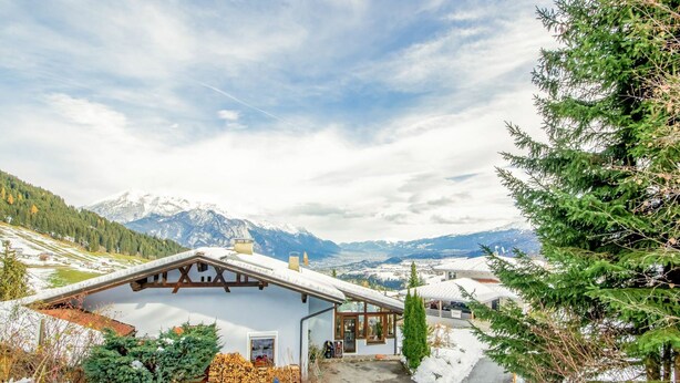 Ein Chalet mit schneebedecktem Dach, einer Holzterrasse und Blick auf verschneite Berge im Hintergrund.