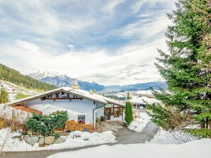 Ein Chalet mit schneebedecktem Dach, einer Holzterrasse und Blick auf verschneite Berge im Hintergrund.