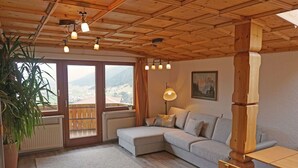 Ein gemütliches Zimmer mit Holzbalkendecke, einem grauen Sofa, einem Holztisch und Blick auf eine gebirgige Landschaft durch große Fenster.