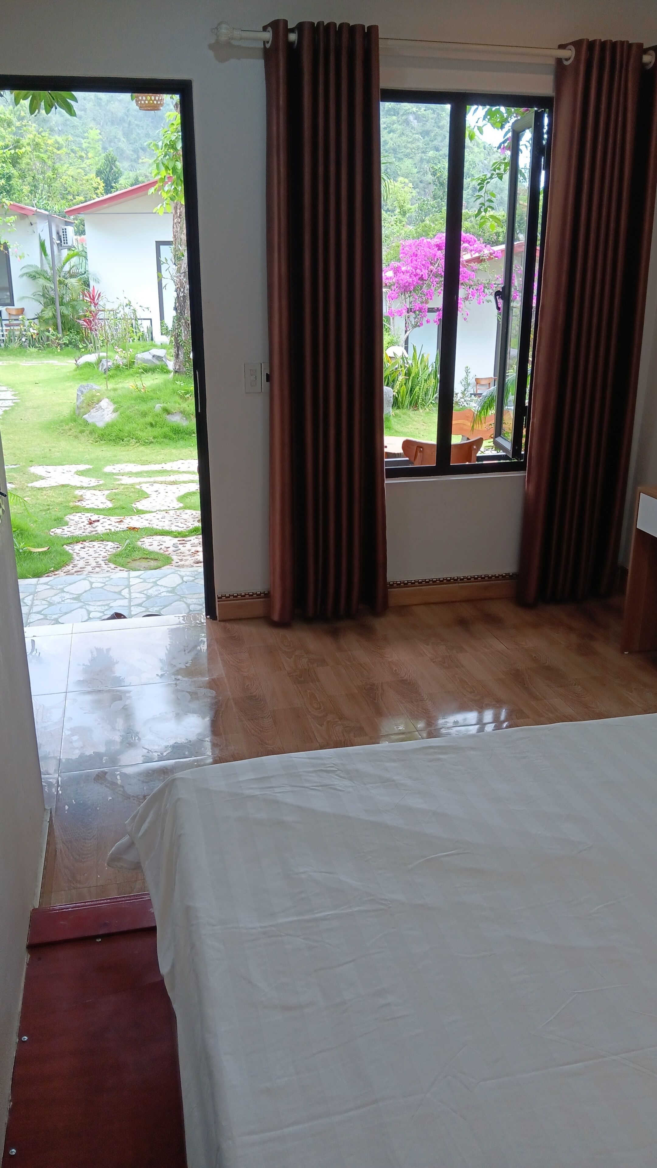 Una habitación con cama, cortinas y vista a un jardín con un edificio de techo rojo.