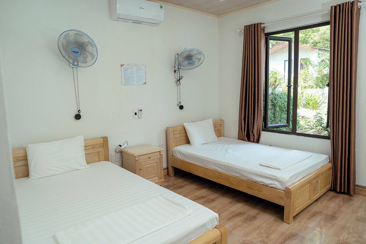 Une chambre d’hôtel avec deux lits simples, une petite table de chevet, un climatiseur fixé au mur, deux ventilateurs muraux, une fenêtre avec des rideaux et une vue sur la verdure environnante.