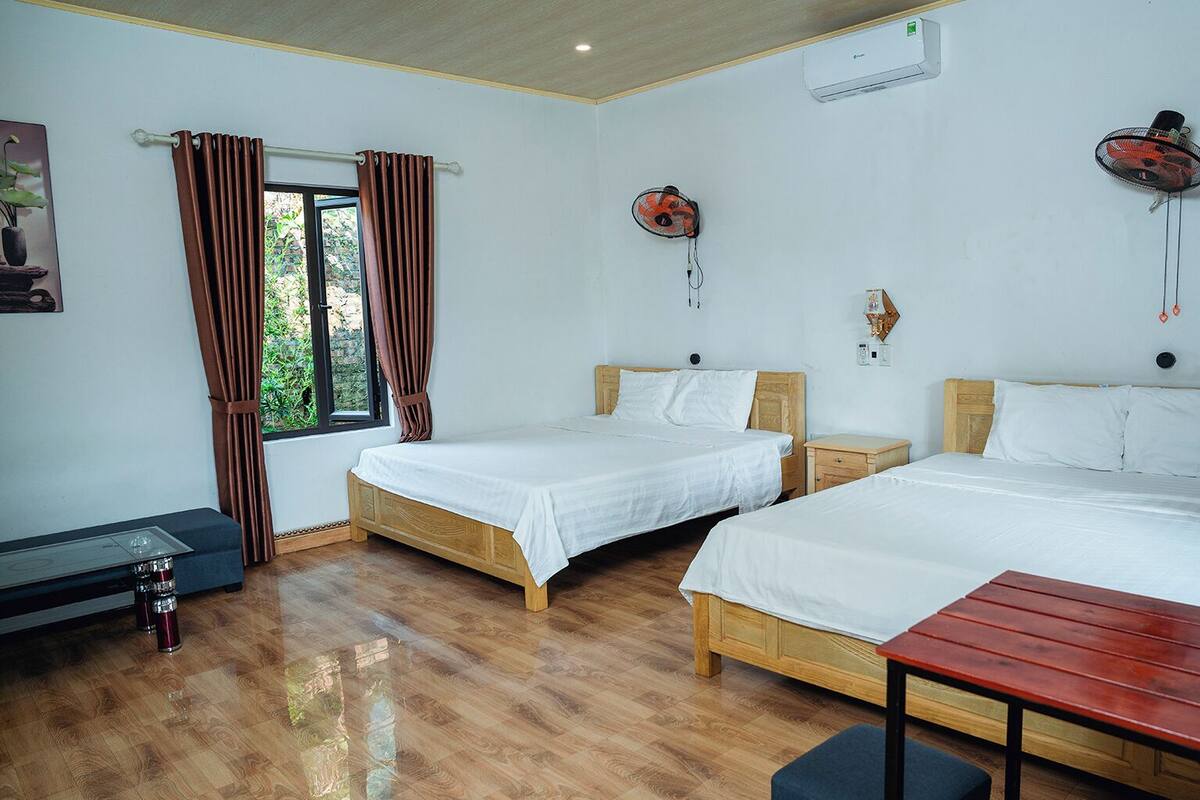 Une chambre d’hôtel avec deux lits en bois, une table en bois, un banc, une fenêtre avec des rideaux, un climatiseur et un ventilateur fixé au mur.