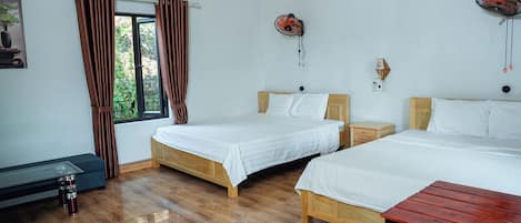 A hotel room with two wooden beds, a wooden table, a bench, a window with curtains, an air conditioning unit, and a wall-mounted fan.