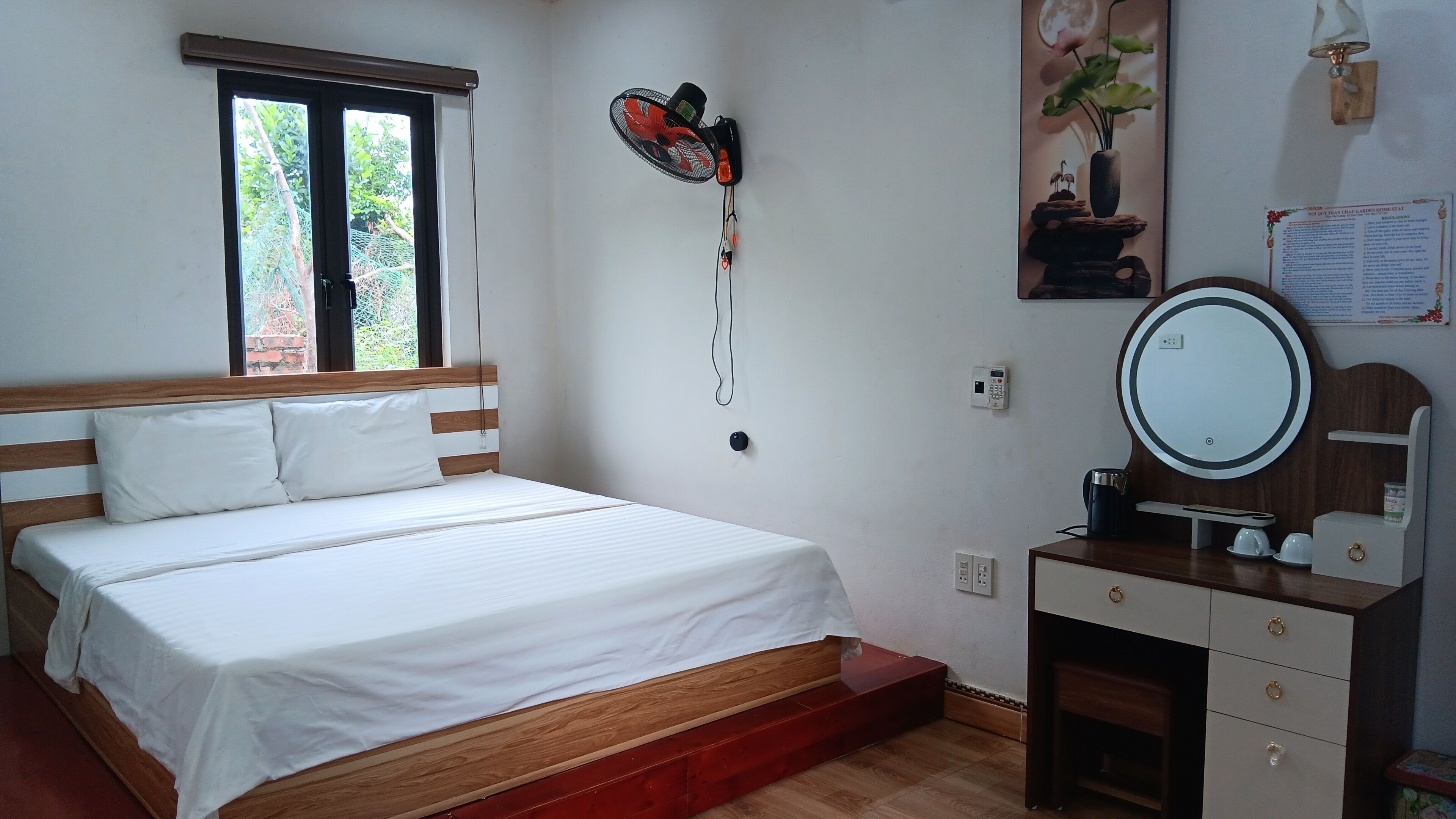 Un dormitorio con cama, mesita de noche con espejo, ventilador de pared y ventana con vistas a zonas verdes.