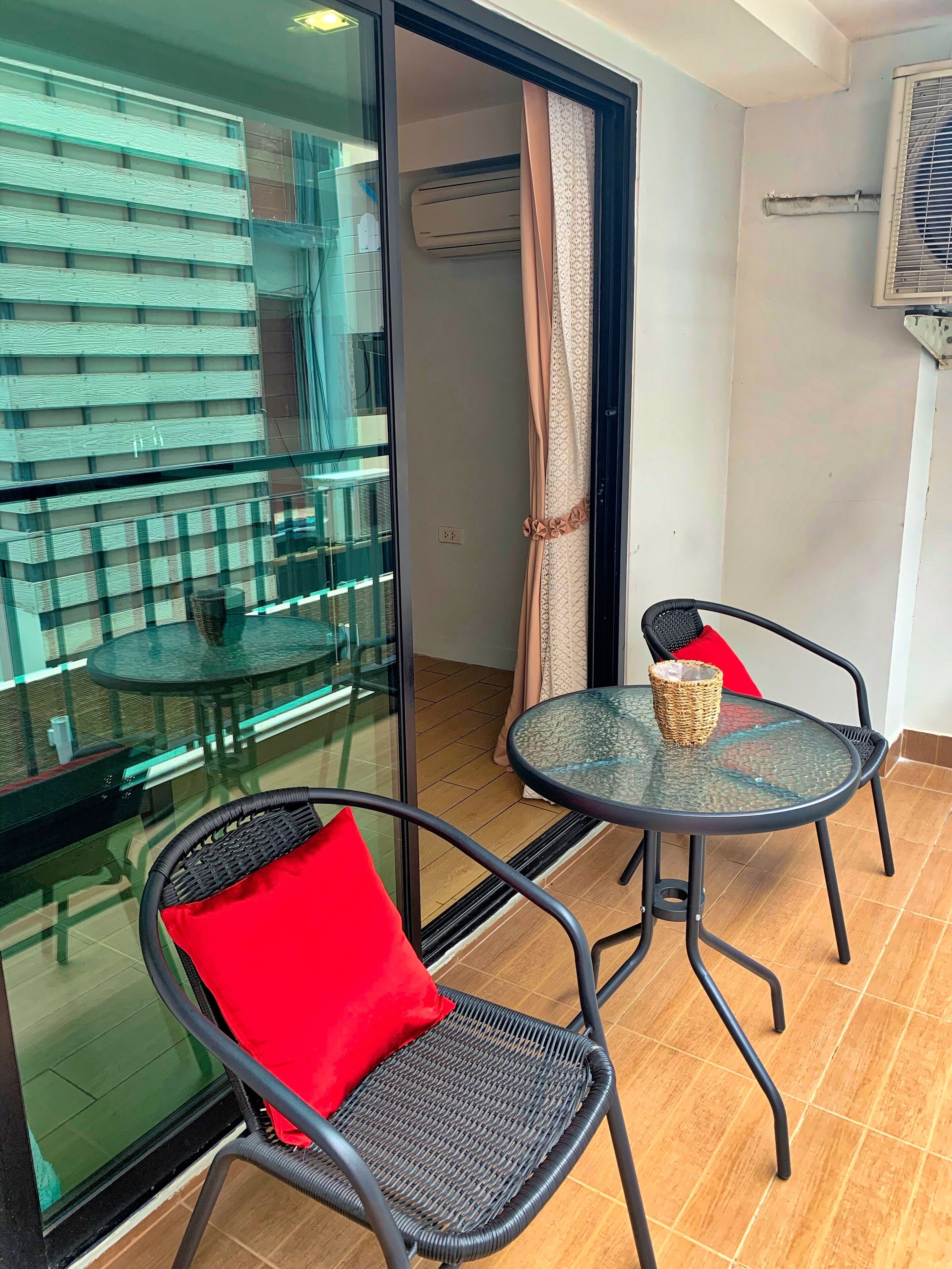 A balcony with a glass door, two wicker chairs with red cushions, a glass table with a woven basket, and an air conditioning unit on the wall.