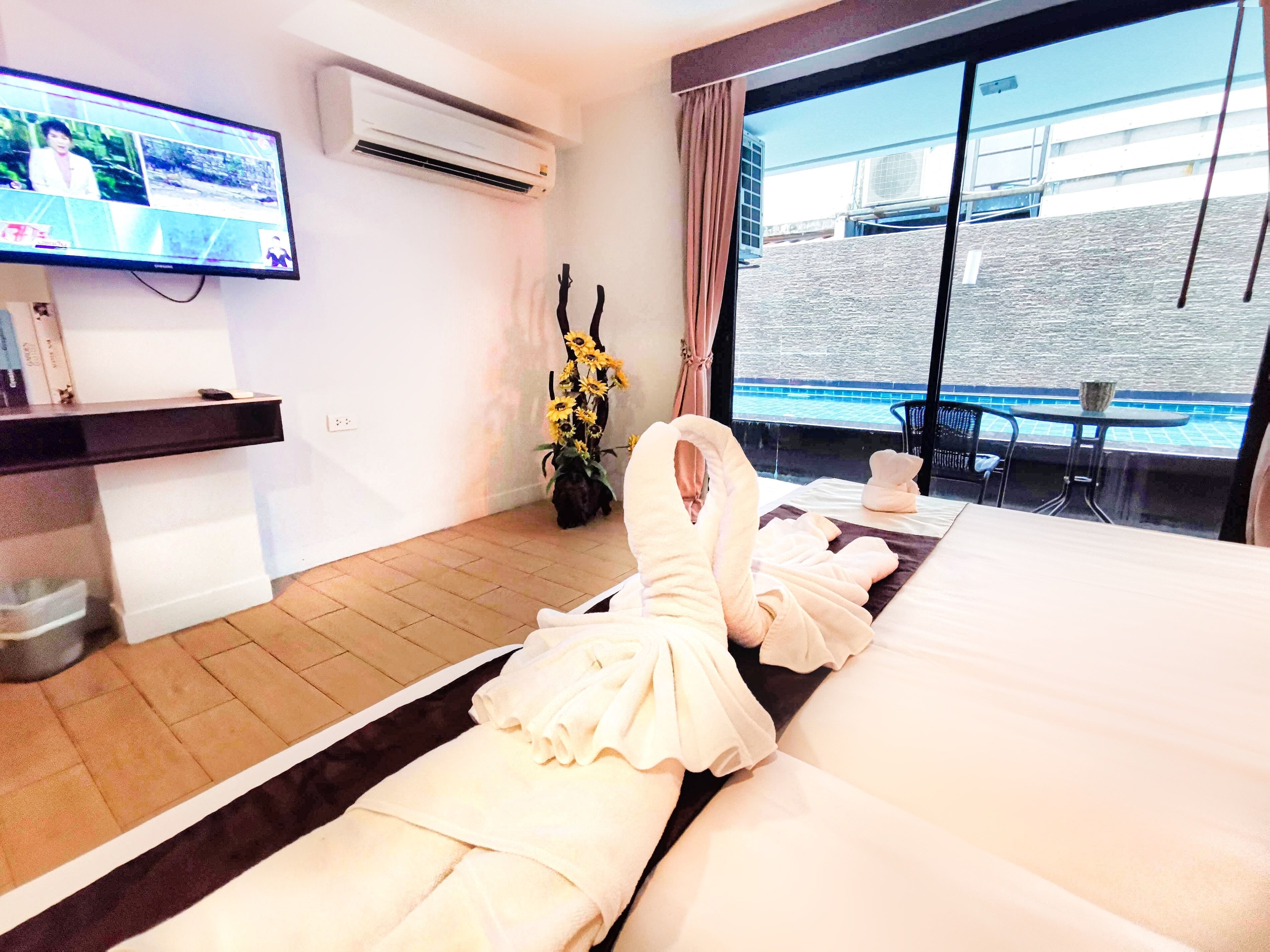 Habitación de hotel con una cama grande, un televisor montado en la pared, un equipo de aire acondicionado, una ventana con vistas a un edificio y una piscina, y una pequeña mesa con una silla.