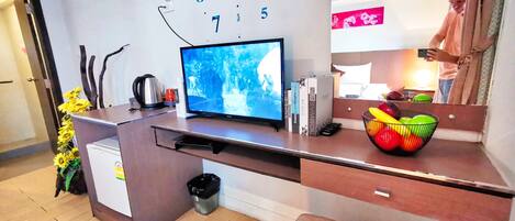 A modern hotel room with a wooden desk, a flat-screen TV, a coffee maker, and a fruit bowl.