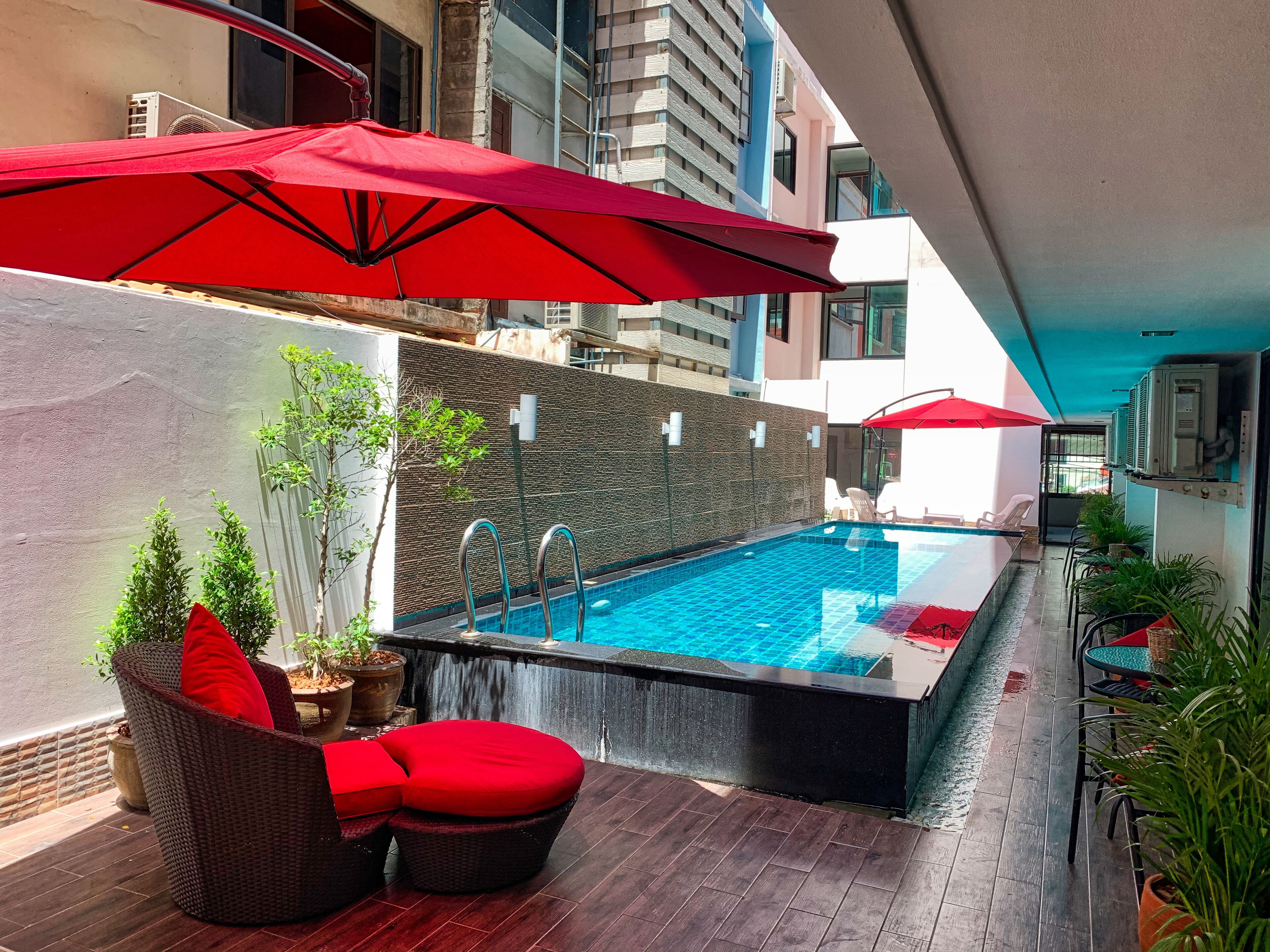 A rooftop pool area with a small pool, red umbrellas, wicker furniture, and potted plants.