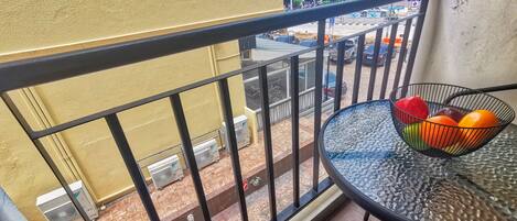 A balcony with a metal railing, a table with a wire basket containing fruits, and a view of the street below.
