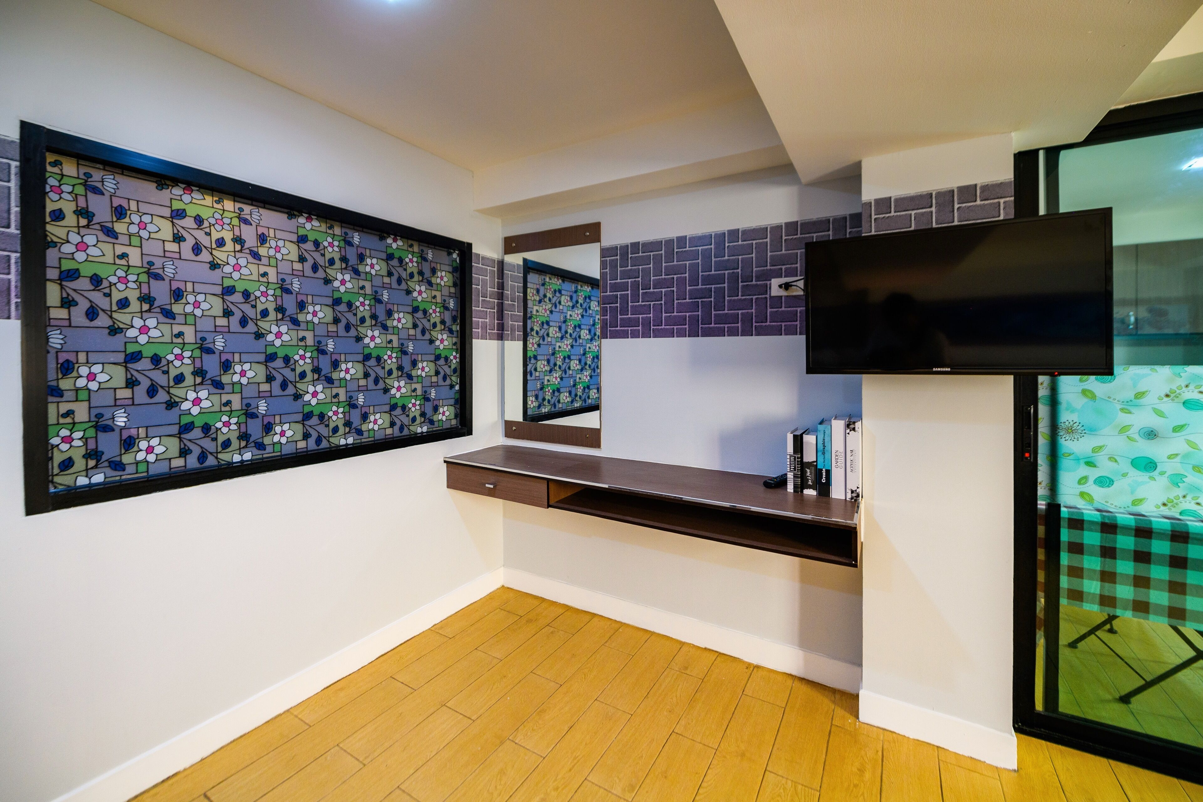 Une pièce avec un plancher en bois, un téléviseur fixé au mur, un bureau avec un miroir et un panneau décoratif mural.