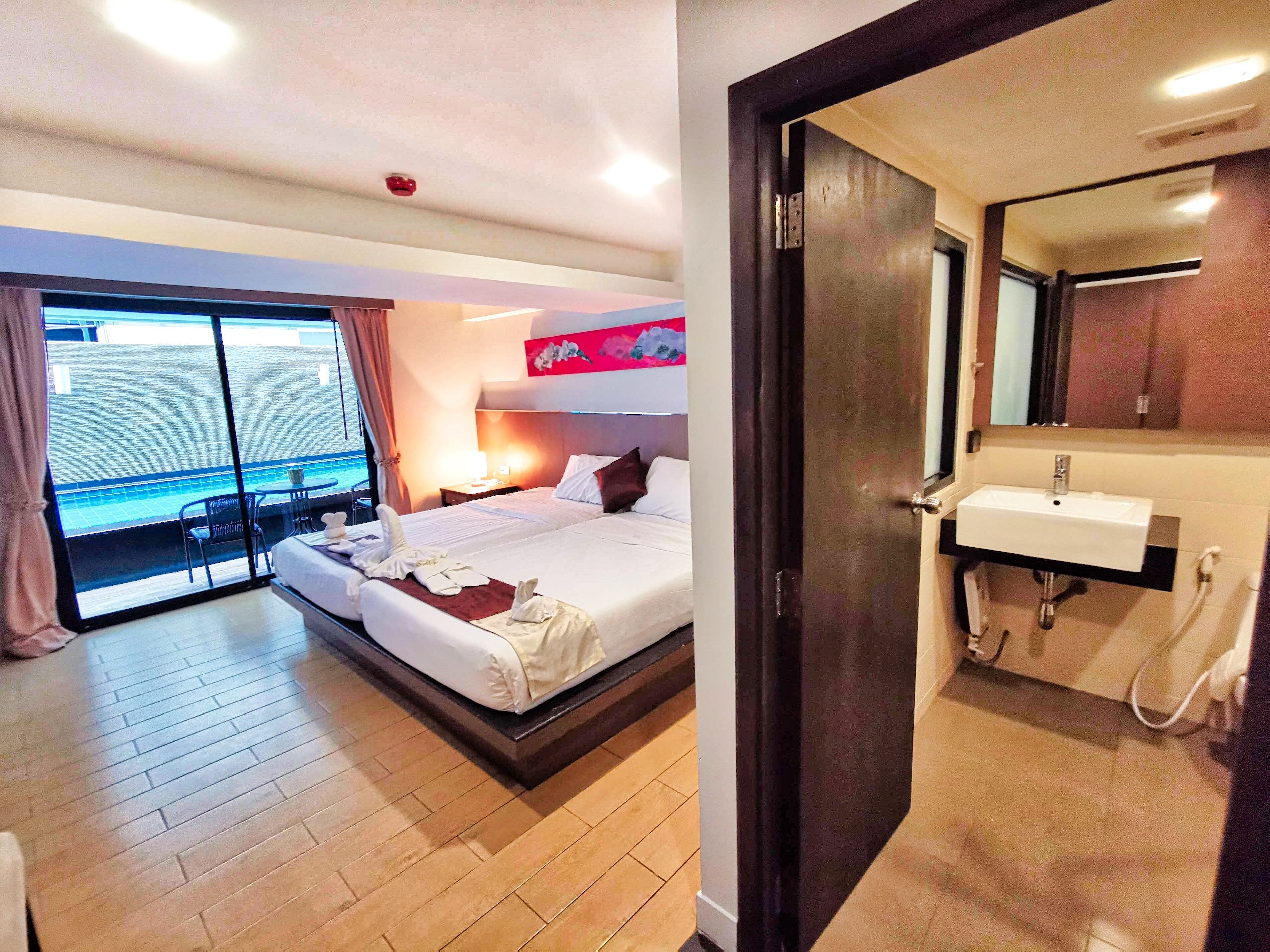 A hotel room with a bed, a bedside table, a window with a view of the sea, and a bathroom with a sink and mirror.
