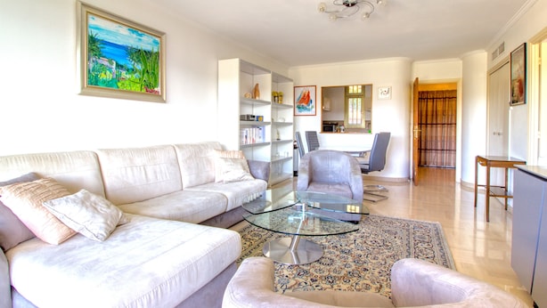 Ein Wohnzimmer mit einer Couch, einem Sessel und einem Glastisch. Im Hintergrund ist eine Küche mit Essbereich zu sehen.