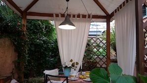 Un espacio al aire libre cubierto con zona de estar y una mesa preparada para el té, rodeado de abundante vegetación y una estructura de pérgola de madera.