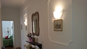 A hallway with a white wall, a mirror, a framed picture, and a shelf with a vase of flowers.