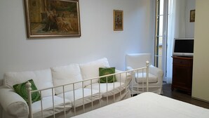 A bedroom with a white bed, a white sofa with green pillows, a small white chair, a painting on the wall, and a television on a wooden stand. - Casa Vicki (Genoa)