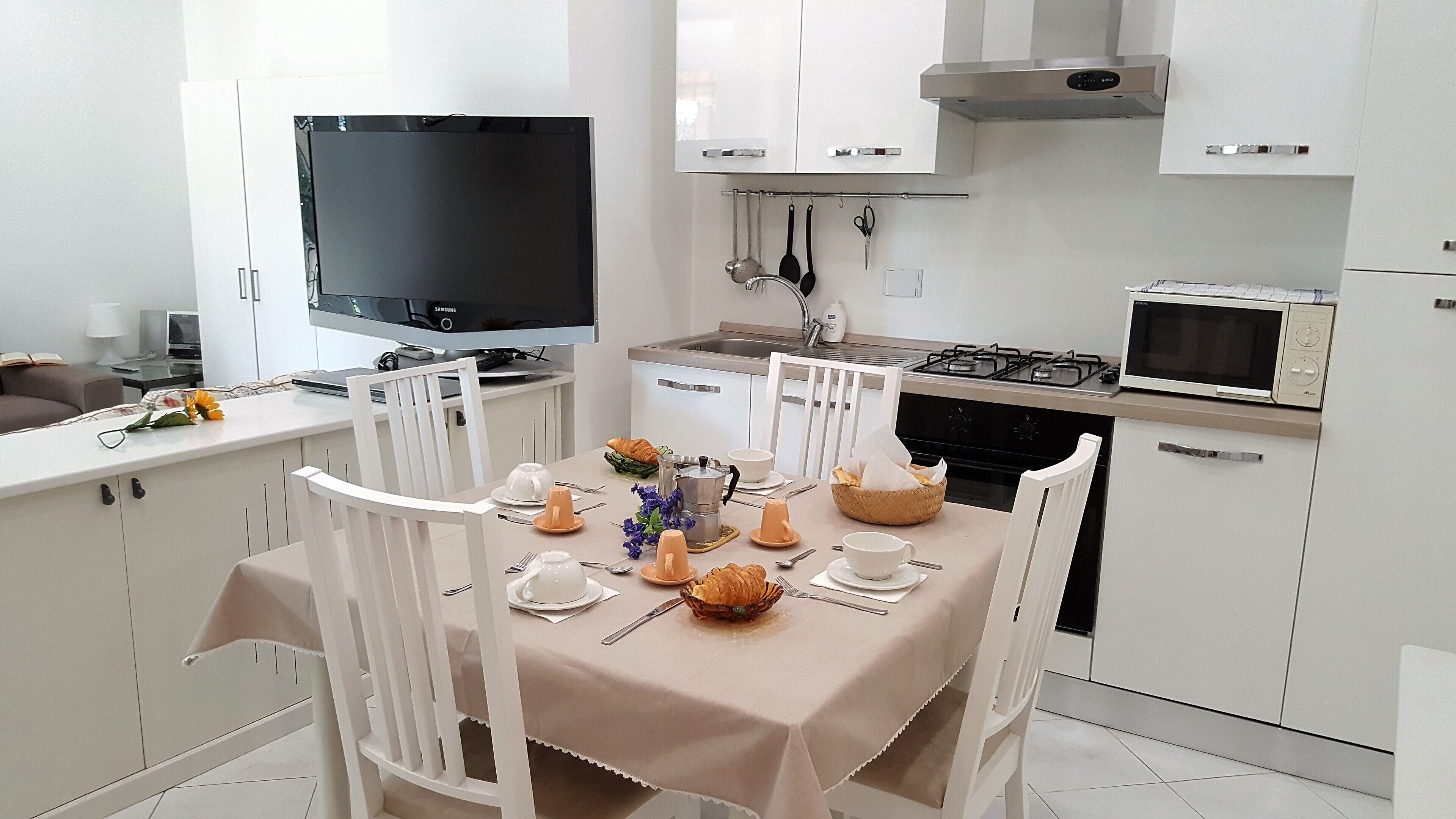 A modern kitchen with white cabinetry, a dining table set for four, and a flat-screen TV above the counter.