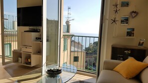 Un soggiorno con tavolo in vetro, divano e televisore. È presente un balcone con vista sul mare e sugli edifici.