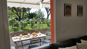 A dining area with a table set for two, chairs, and a view of a garden with trees and a stone wall. - Casa Agrumeto (Genoa)