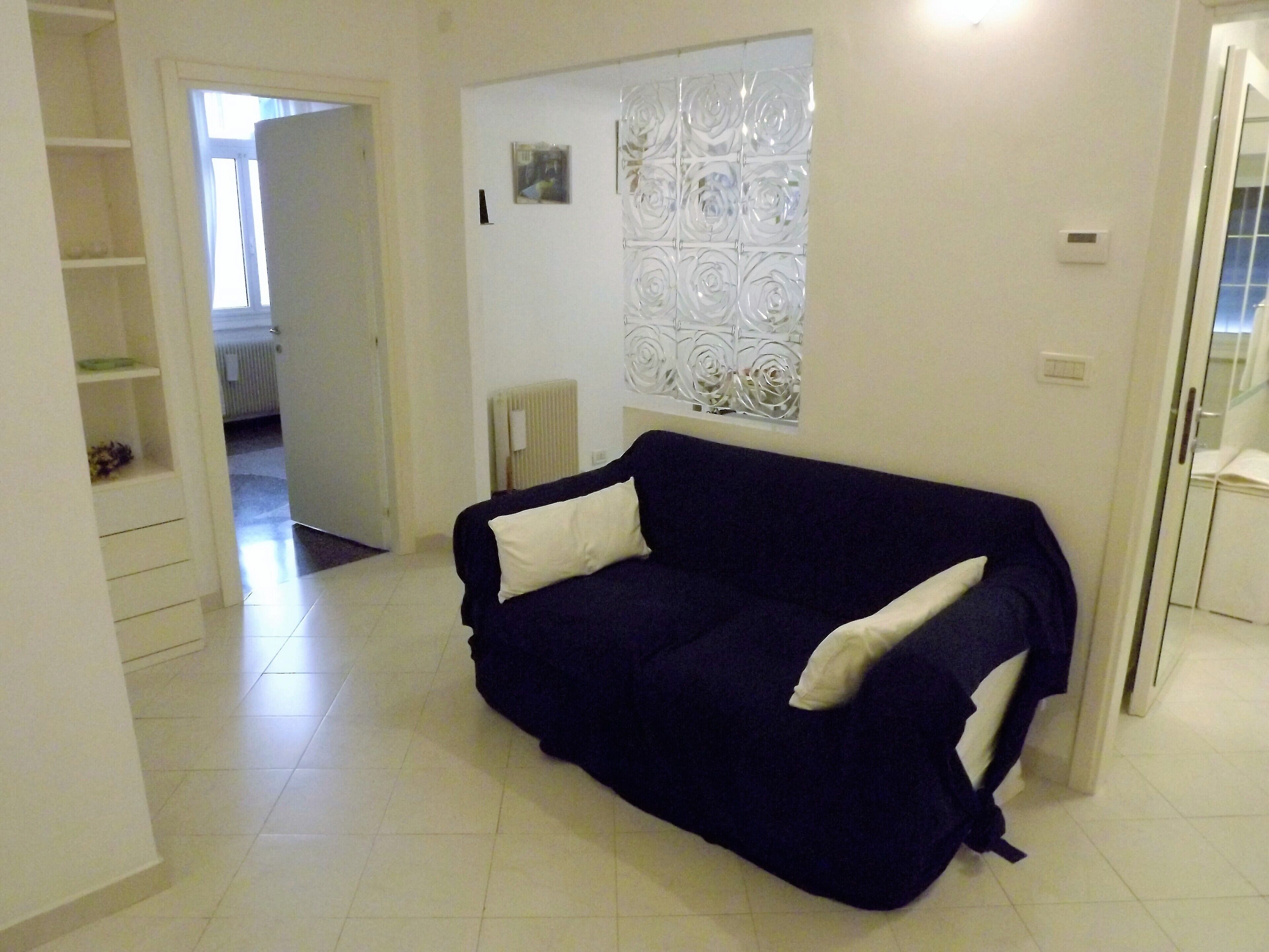 A living room with a dark blue sofa, white cushions, a radiator, a window with decorative glass, and a white door leading to another room.