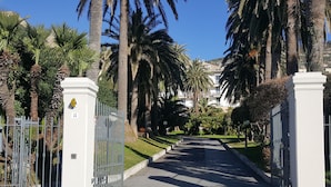 A gated entrance with a pathway leading to a building, surrounded by palm trees and greenery. - House Sole (Genoa)