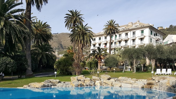 A swimming pool with clear blue water, surrounded by rocks and palm trees. In the background, there is a white building with balconies and a hilly landscape. - House Sole (Genoa)