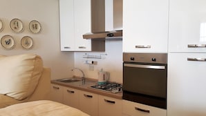 A compact kitchen with a gas stove, sink, and white cabinets. There are decorative plates on the wall and a beige sofa in the living area. - House Betta (Genoa)