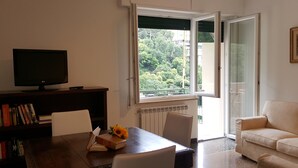 A living room with a TV, books, a table with a book and a flower, a sofa, and a window with a view of greenery. - Casa Barbara (Bogliasco)