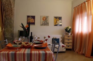 A dining area with a table set for two, featuring a striped tablecloth and various dishes and glasses. There are three framed artworks on the wall, a potted plant, and a wicker side table. - Casa Bussana (Sanremo)