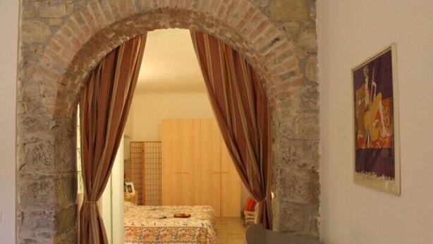 Una habitación con un arco de piedra que da a una cama con cubrecama floral. Una silla y una mesita se ven al frente.