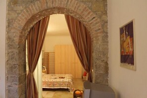 A room with a stone archway leading to a bed with a floral bedspread. A chair and a small table are visible in the foreground. - Casa Bussana (Sanremo)