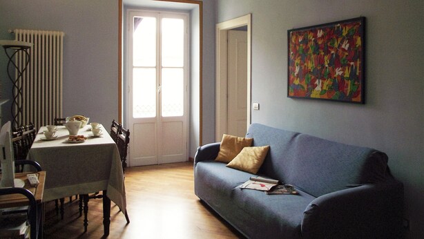 A living room with a blue sofa, a dining table with tea cups and a bowl, a radiator, a door, and a colorful painting on the wall. - Azzurro Pervinca (Finale Ligure)