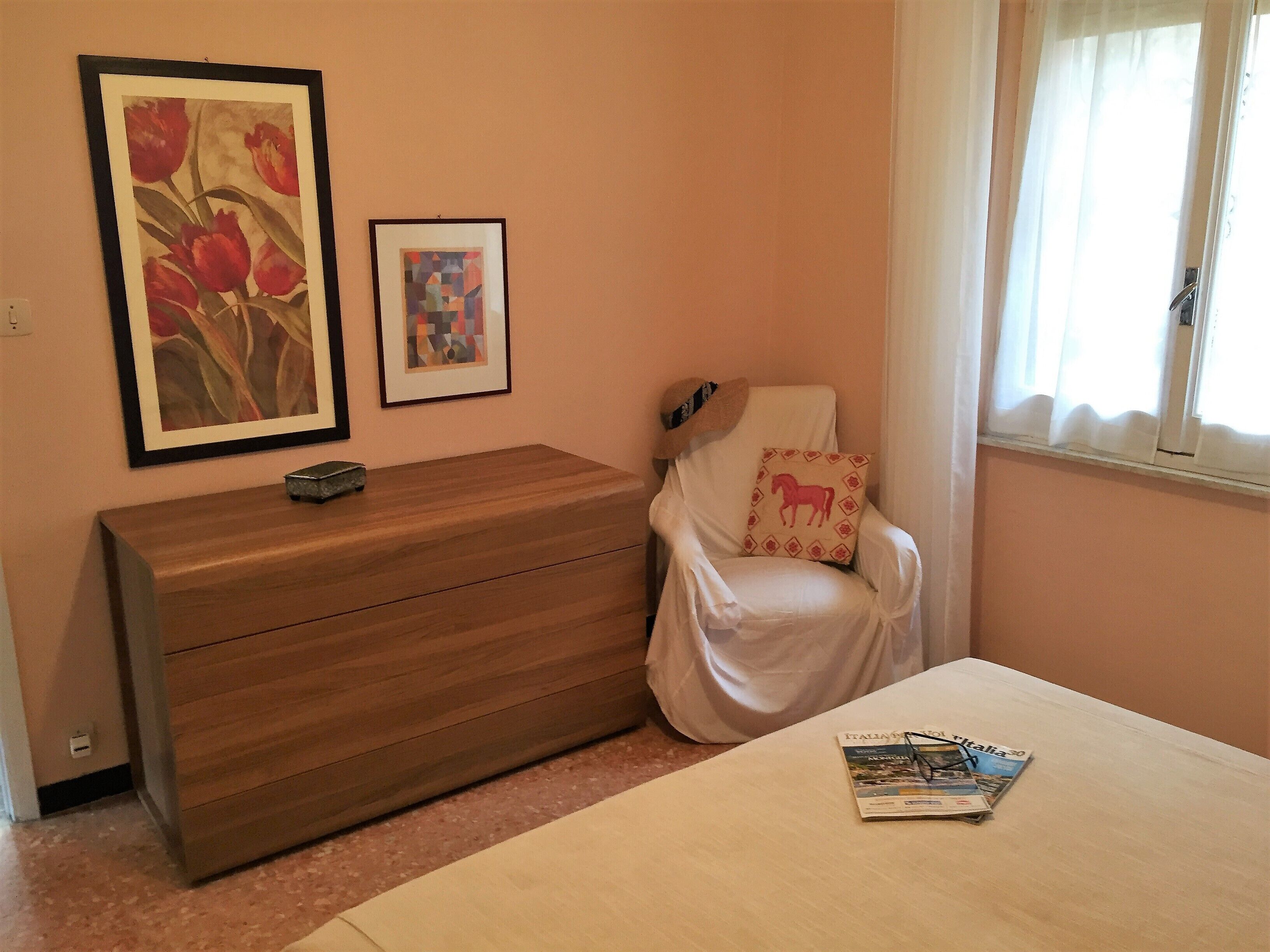Une chambre à coucher avec une commode en bois, une chaise agrémentée d’un coussin décoratif, un tableau encadré et un magazine posé sur le lit.