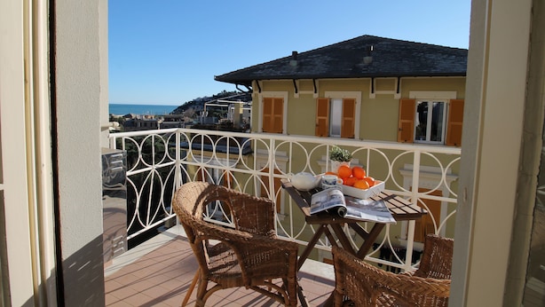 A balcony with wicker furniture, a table with a newspaper and fruit, and a view of the sea and buildings. - House Bella (Arenzano)